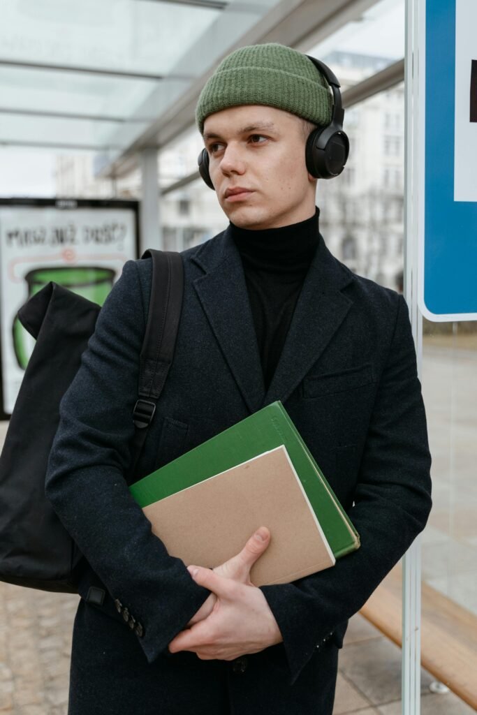 You man wearing a fitted knit winter hat, over-the-ear headphones, and coat, with    a bookbag slung over one shoulder to demonstrate a comfortable winter commuter